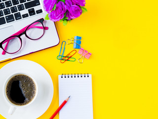 Flat lay, top view office table desk frame. office accessories including calculator, mouse laptop, glasses, clips, red pencil, black coffee, note book, blue pen and pink flower on yellow background.