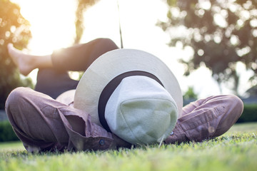 Asian trendy hipster man wearing  a white cap, pink shirt and black jean while lying on the green grass for relaxing on at park in the city with blurry tree background and lens flare