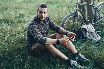 Wall Mural - Handsome Young Man Cyclist Sits On Summer Meadow Near Bicycle, Holding At Tablet Recreation Resting Travel Destination Concept