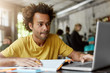 © wayhome.studio  - Portrait of clever student with dark skin and bushy hair wearing casual clothes while sitting at cafeteria working at his course paper searching for information in internet using his laptop computer