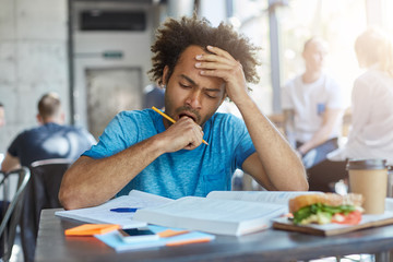 Wall Mural - Male student with fine crop of hair and bristle sitting at university canteen drinking coffee and eatting fast food being exhausted after sleepless night of studying yawning while looking in books