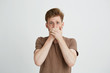© Cookie Studio - Portrait of surprised frightened scared young man looking at camera closing mouth with hands over white background.