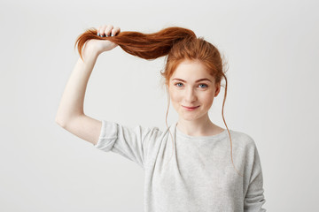 Wall Mural - Portrait of young beautiful redhead girl touching her hair tail looking at camera over white background.