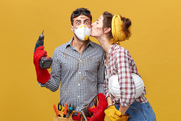 Pretty female kising her husband in cheek being thankful to him for repearing her wardrobe. Surprised male worker in mask holding drilling machine being glad to recieve kiss from his girlfriend