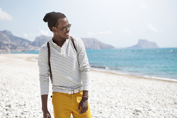 Relaxed black young male in trendy black hat, sunglasses, yellow trousers and casual sweater holding rucksack having walk on seaside looking at blue sea admiring sunny weather during vacations