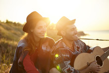 Romantic Man With Guitar Singing Serenade To His Girlfriend Oudoors. Lovely Couple In Love Sitting Together At Greenland Enjoying Beautiful Sunset Looking Into Distance With Smile And Happiness.