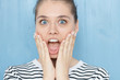 Cute young caucasian female in navy striped t-shirt with opening mouth widely, saying omg, wow, having excited astonished look, holding hands on her face standing against on blue background