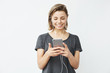 © Cookie Studio - Young beautiful girl in headphones smiling looking at phone screen over white background.