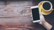 © Farknot Architect - Mockup image of hands holding white mobile phone with blank black screen with yellow . hot coffee cups on vintage wooden table in restaurant