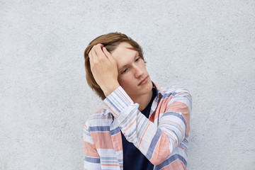 Portrait of good-looking male with trendy hairstyle holding his hand on hair looking aside with dreamy expression. Teenage boy posing against white wall dressed casually thinking about something