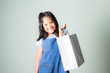 pretty  little asian girl in blue jeans dungarees holding shopping bags with white concrete wall background.portrait of young kid smiling and looking at camera