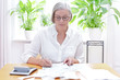 Deutsche Seniorin füllt die Formulare für die jährliche Steuererklärung aus, im Wohnzimmer an einem Tisch mit Taschenrechner und vielen Rechnungen und Quittungen.