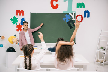 Wall Mural - Schoolboy holding chalk and pointing at blackboard while classmates raising hands