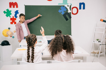 Wall Mural - Back view of classmates looking at schoolboy answering near blank chalkboard