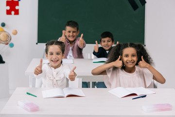 Wall Mural - Cheerful multiethnic schoolkids showing thumbs up while sitting at desks in classroom