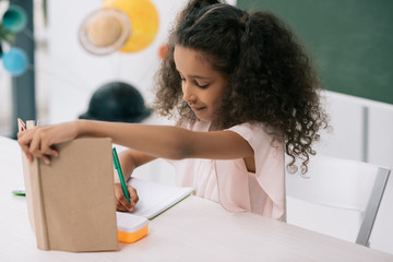 Wall Mural - Focused african american schoolgirl holding textbook and writing exercise at lesson