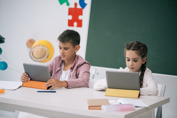 Wall Mural - Concentrated schoolboy and schoolgirl using digital tablets while sitting at desk in classroom