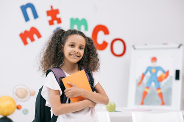 Wall Mural - Adorable african american schoolgirl with backpack holding textbook and smiling at camera in classroom