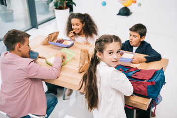 Wall Mural - Multiethnic group of schoolkids eating lunch while sitting together at table