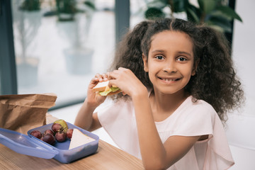 Wall Mural - Cute african american schoolgirl eating sandwich and smiling at camera at lunch in school