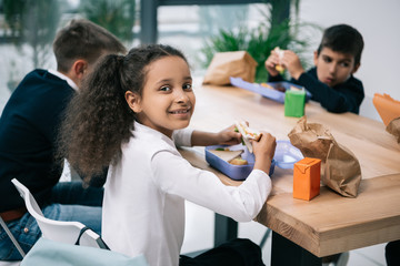 Wall Mural - Cute multiethnic schoolkids eating lunch while sitting at table