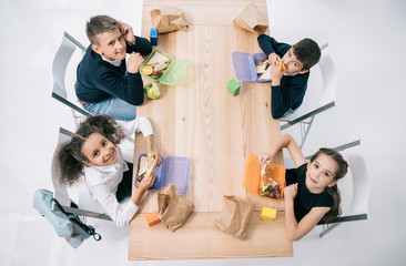 Wall Mural - Overhead view of multiethnic schoolkids eating lunch while sitting at table and smiling at camera