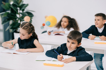 Wall Mural - Cute little schoolchildren sitting at desks and writing in exercise books