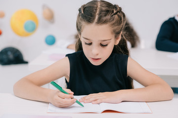 Wall Mural - Close-up portrait of cute concentrated schoolgirl writing in exercise book at lesson