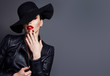 Beautiful young mysterious girl in a black hat and black leather jacket on a gray background. Eyes are covered with a hat. Makeup - red lips. Manicure - long red nails, nail polish. Fashion, beauty.