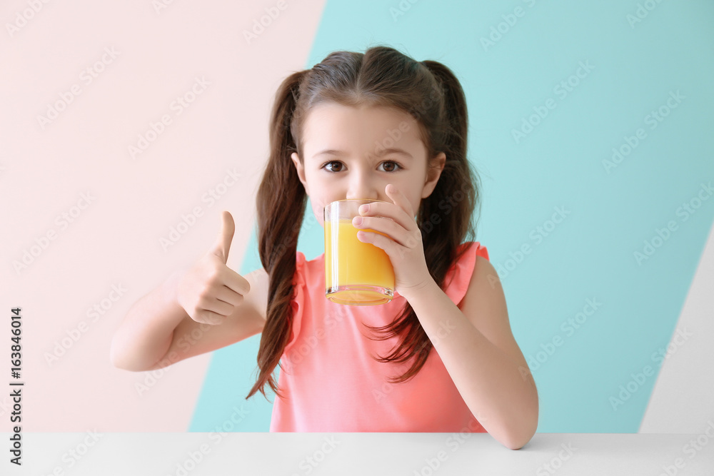 Cute little girl drinking juice while sitting at table, on color background