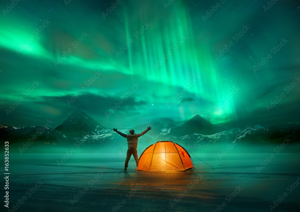 A man camping in wild northern mountains with an illuminated tent viewing a spectacular green northern lights aurora display. Photo composition. - obrazy, fototapety, plakaty 