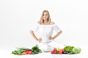 Wall Mural - Beautiful upset blond woman in white clothes and lots of fresh vegetables on white background. diet