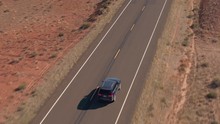 AERIAL DISTANCING: Flying Above A Black SUV Car Driving Along An Empty Road Trough Sandstone Desert In Utah Flatlands, USA. People On Road Trip Traveling On Road Trough Desert Landscape On Sunny Day.
