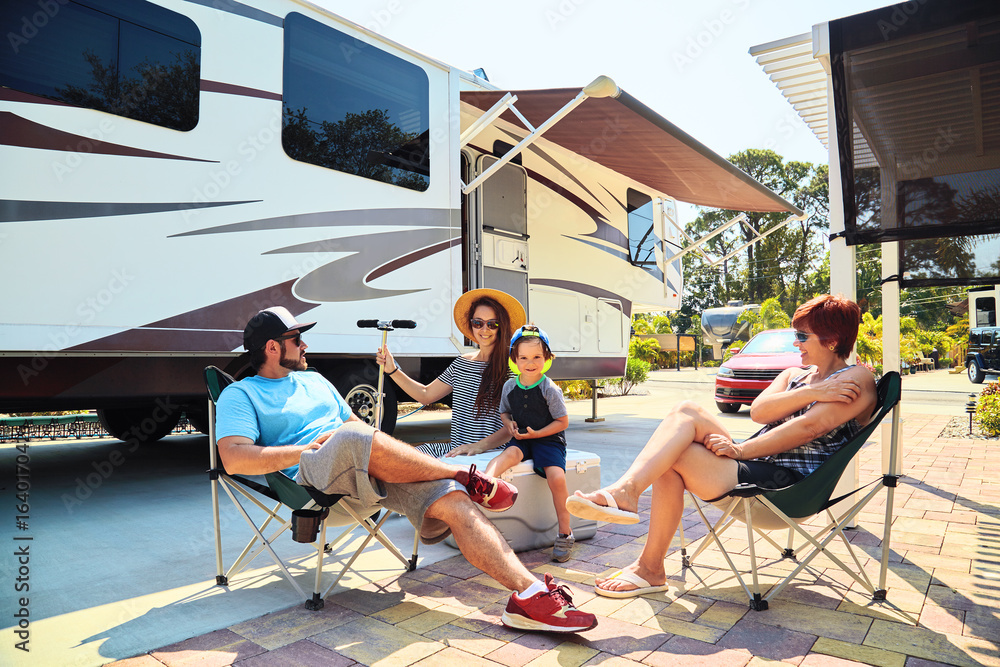 Mother,father,son and grandmother sitting near camping trailer,smiling.Woman,men,kid relaxing on chairs near car.Family spending time together on vacation near sea or ocean in modern rv park - obrazy, fototapety, plakaty 
