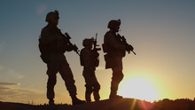 Squad Of Three Fully Equipped And Armed Soldiers Standing On Hill In Desert Environment In Sunset Light.