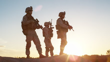 Squad Of Three Fully Equipped And Armed Soldiers Standing On Hill In Desert Environment In Sunset Light.
