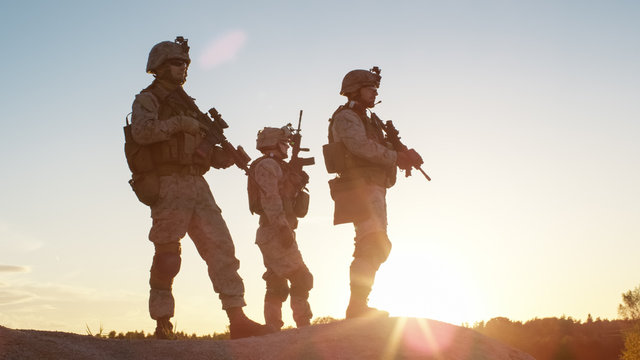 squad of three fully equipped and armed soldiers standing on hill in desert environment in sunset li