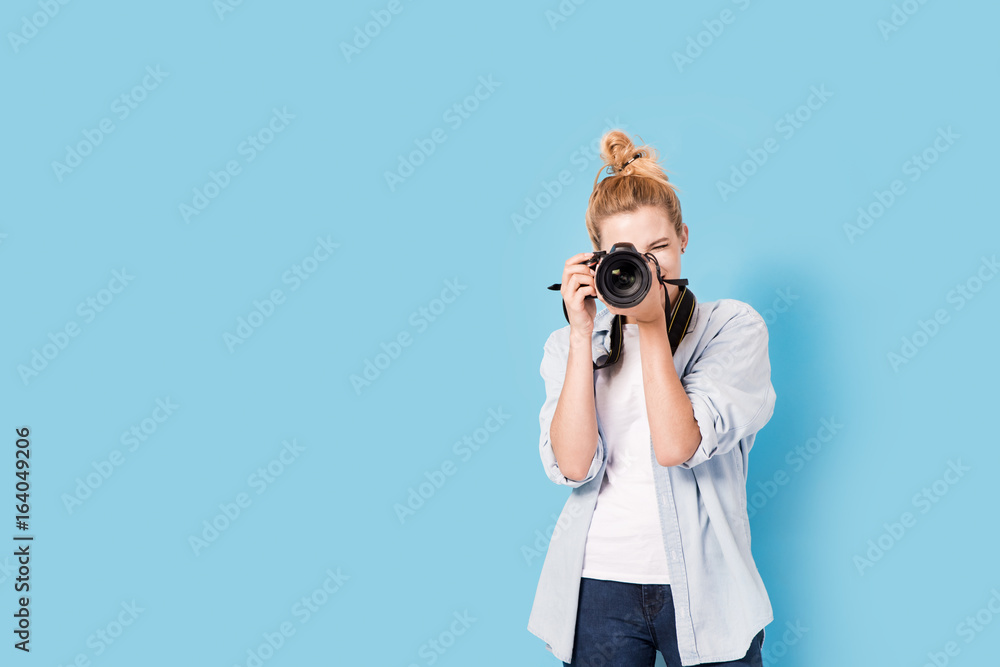 Young blonde photographer is taking a photo. Model isolated on a blue background with copy space - obrazy, fototapety, plakaty 