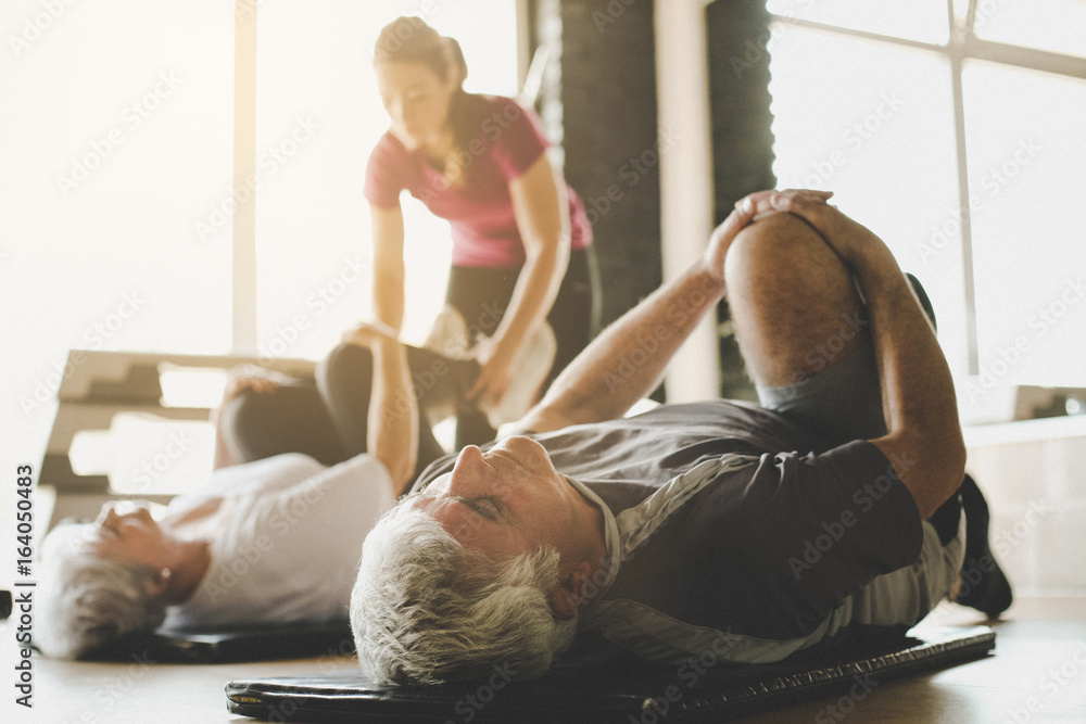 Senior couple workout in rehabilitation center. Personal trainer helps elderly couple to do stretching on the floor. Focus on man.