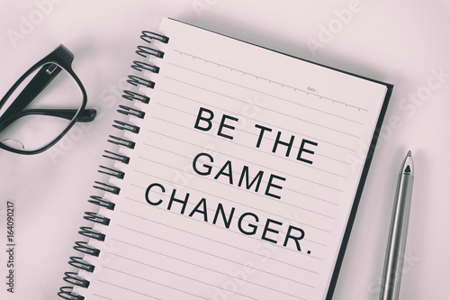 Fotografie, Obraz Inspirational quote - be the game changer: written on a note pad with eyeglasses and pen