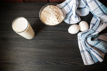 The process of preparation in detail. Cooking, kneading dough. A glass of milk, 2 eggs, a saucer with flour and a blue napkin on a black wooden table