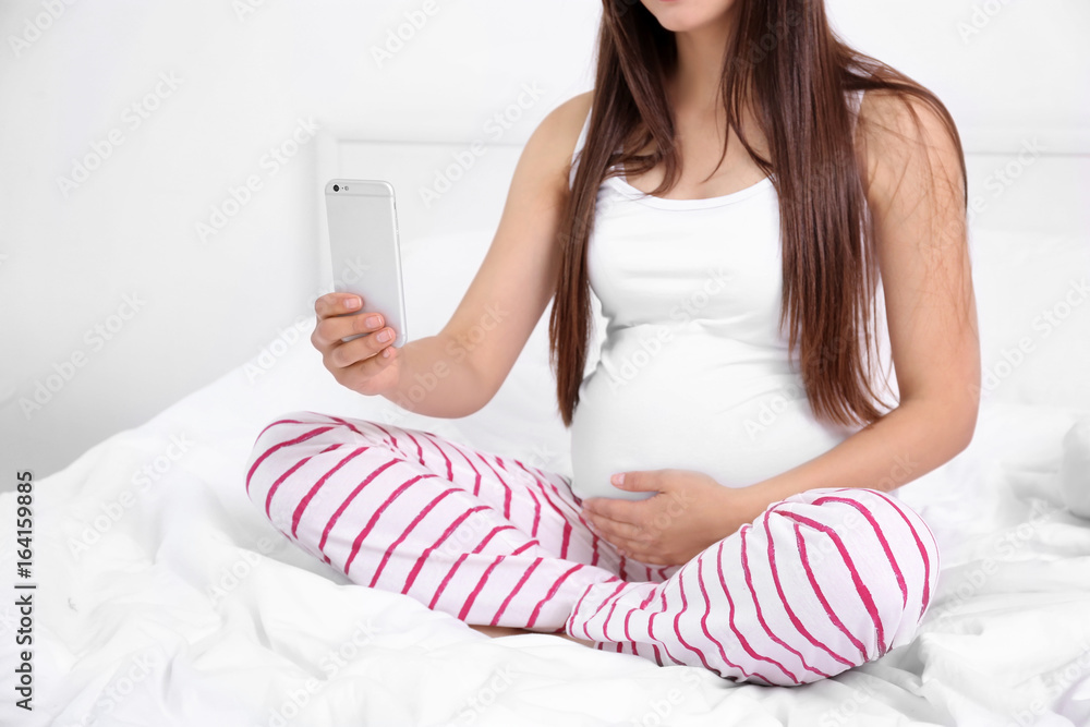 Pregnant woman with mobile phone sitting on bed