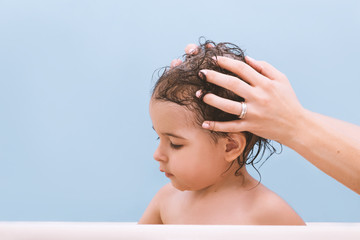 Cute happy toddler baby taking a bath. Mother washing hair to her daughter toddler . Little child in a bathtub. Smiling kid in bathroom on blue background. Infant bathing. Hygiene and health care.