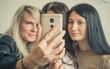 © Artem Markin - Three beautiful young cute girlfriends make selfie, photo on yellow phone, smiling and laughing in office during lunch break. Girls with black hair and blond girl