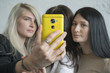 © Artem Markin - Three beautiful young cute girlfriends make selfie, photo on yellow phone, smiling and laughing in office during lunch break. Girls with black hair and blond girl