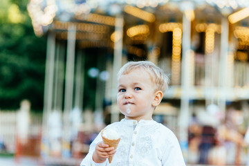 Funny toddler boy in white shirt and blue pants eating an ice cream in front of the carousel