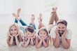 © deagreez - High angle view of happy family of four relatives, lying with crossed legs on the beige carpet on the floor, holding heads with arms, smiling, at home