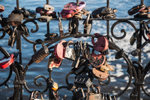 A Lot Of Wedding Padlocks Of Different Sizes And Shapes On The Curved Cast-iron Black Grate On The Background Of Blue Water