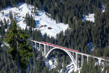 Horizontal View Of A Red Train Crossing A Bridge Over A Deep Gorge In The Swiss Alps Near Arosa And Chur In Winter