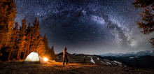 Male Tourist Have A Rest In His Camp Near The Forest At Night. Man Standing Near Campfire And Tent Under Beautiful Night Sky Full Of Stars And Milky Way, And Enjoying Night Scene. Panoramic Landscape